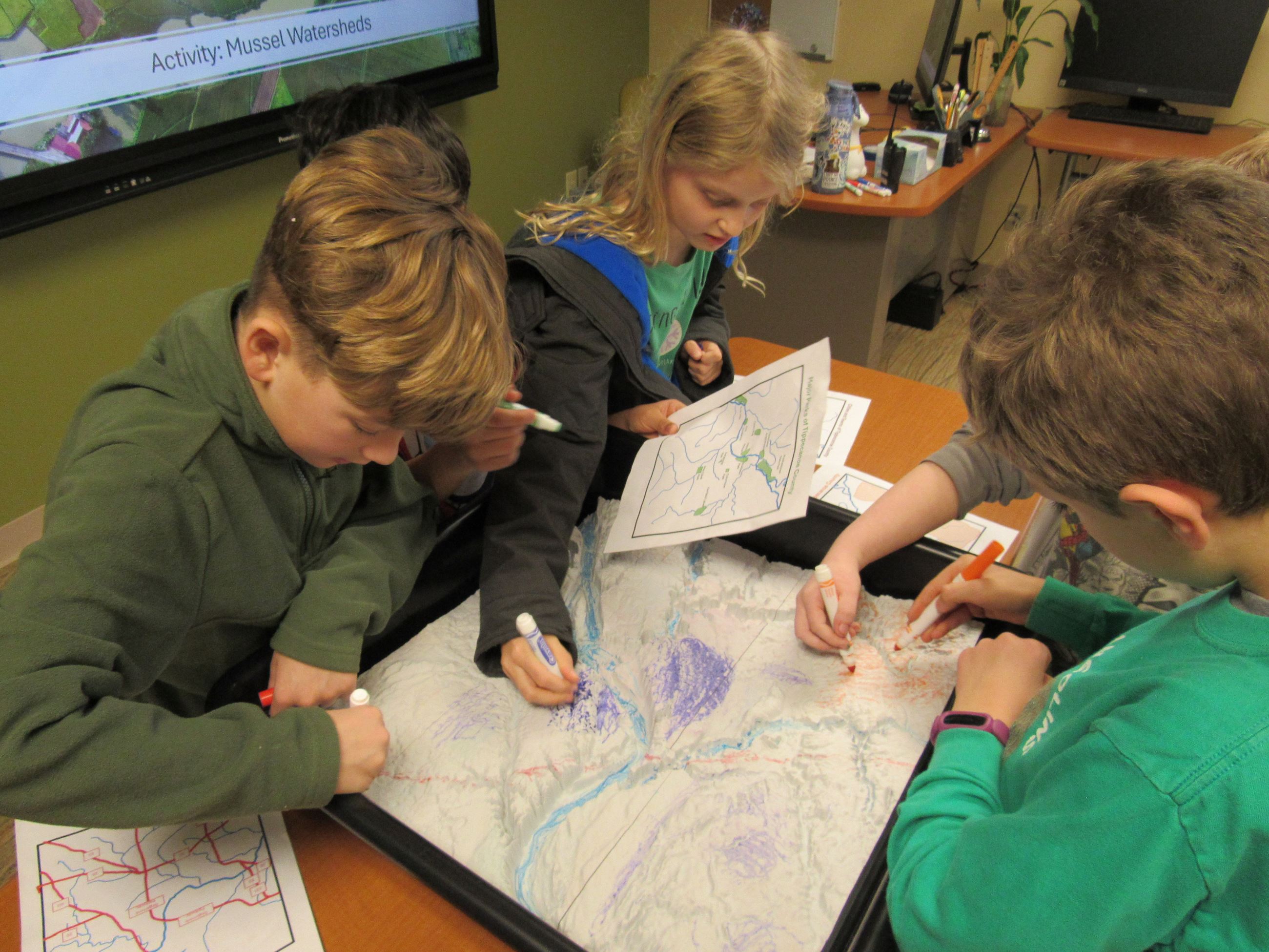 Image of children using paper maps to draw landmarks on a 3d topographic map.